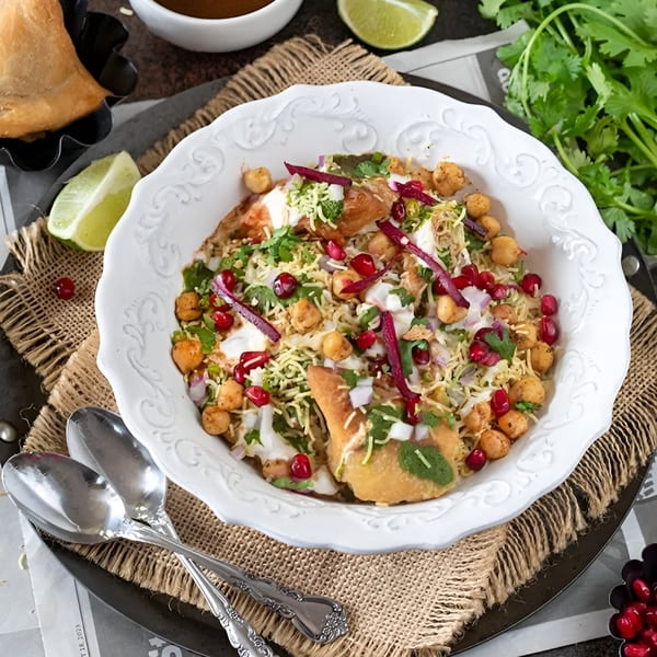 A white bowl of Indian samosa chaat, garnished with pomegranate, chickpeas, and fresh herbs.