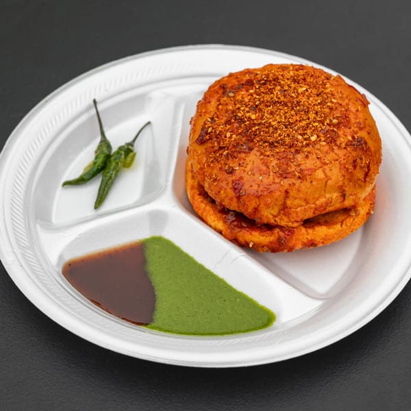 Bombay street food: a vada pav with green chilies and two chutneys on a white divided plate.