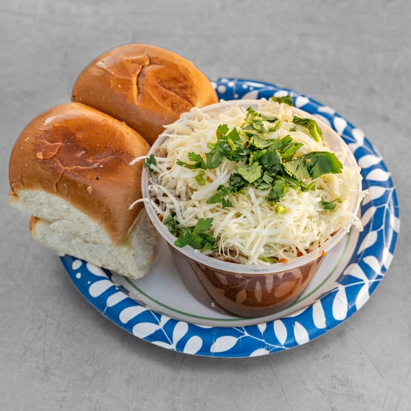 Pav Bhaji, a Bombay street food, with two bread rolls and a bowl of vegetable curry topped with cheese and cilantro.