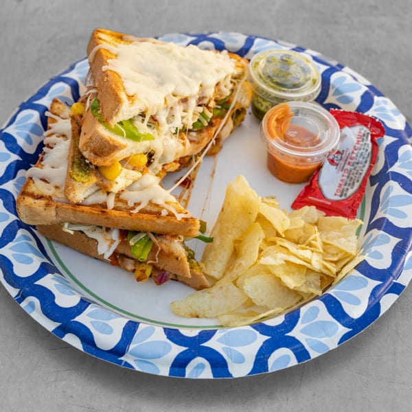 Grilled Bombay street food sandwich with melted cheese, chips, and dipping sauces on a paper plate.