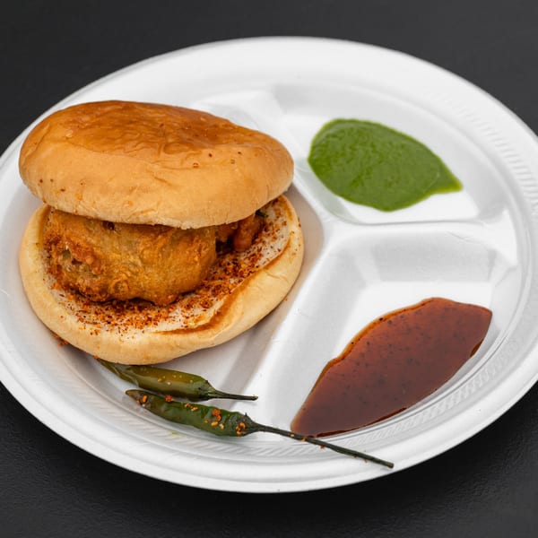 A vada pav, green chutney, and tamarind chutney on a white partitioned plate with green chilies.